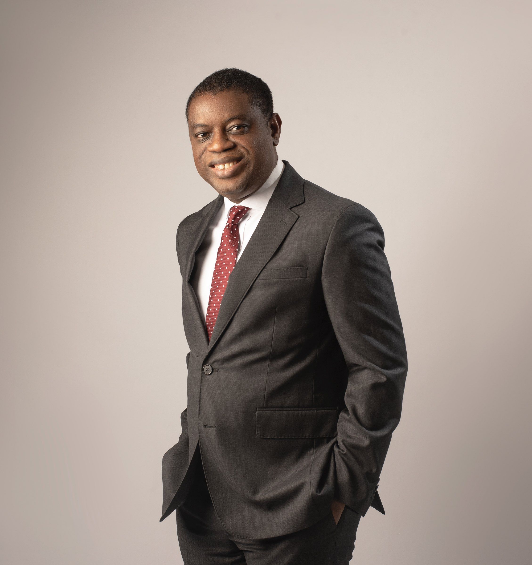 Akin Monehin in a dark suit and red tie, standing against a plain grey background.