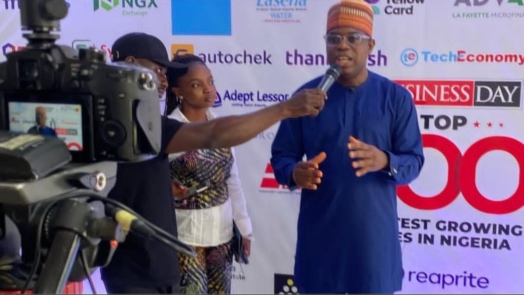Akin Monehin in a blue kaftan and red cap answering a reporter’s questions in front of a television camera, a BusinessDay backdrop behind him.